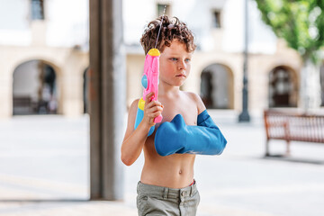 child has fun with a water pistol in summer with a rubber band to protect his cast arm