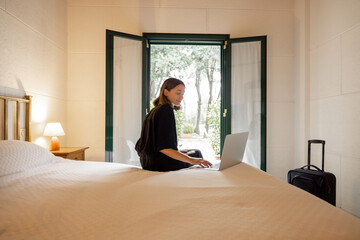Woman lying on bed and using smartphone and laptop computer in hotel room. Concept of weekend, tourism and vacation. Idea of leisure and entertainment. Young caucasian girl resting on resort