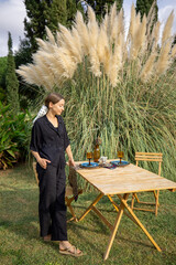 Woman preparing for lunch at garden, standing near the served table and beautiful pampas grass on...