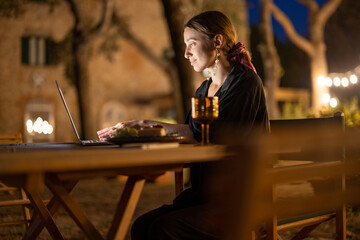 Caucasian woman watching something on laptop computer at evening time outdoors. Young woman sitting at table, eating fruits and drinking wine. Concept of weekend, rest and vacation. Idea of leisure