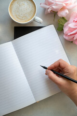 Open note book and female hands holding a pencil. Empty page mockup, cup of coffee, pink roses flowers on bright background. Table top, to do list.