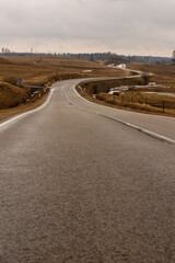 Vertical photo of tarmac road curving through brown meadows at early spring. Life is not a straight road – melancholic curvy drive. Wavy gray asphalt path near Ergli, Latvia in rainy day