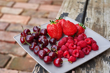 A plate with bright red fruit against the background of a cracked bench and a brick. Summer fruits strawberries raspberries and cherries. Plate of vitamins.