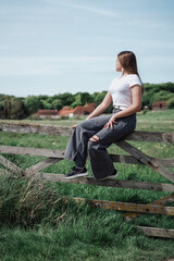 The girl is sitting on a wooden fence in the field. The girl sits on the fence and looks at the green fields.