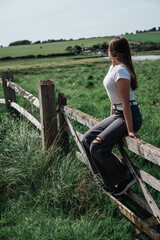 The girl is sitting on a wooden fence in the field. The girl sits on the fence and looks at the green fields.