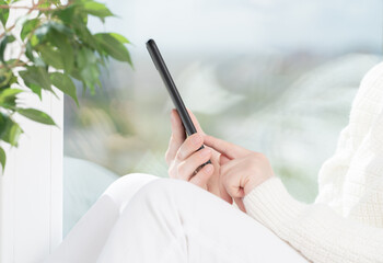 Girl sits by window and uses cell phone, looks at smartphone, reads, shop, communicates with...