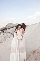 Fototapeta premium female energy practice in desert. two brunette girls in white boho style dresses are standing in hugs on the white sand career background at sunset. lifestyle esoteric concept, free space