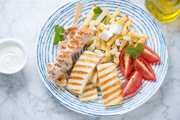 Blue and white plate with grilled halloumi, souvlaki, greek-style fries with feta and tomatoes, studio shot on a light-grey marble background
