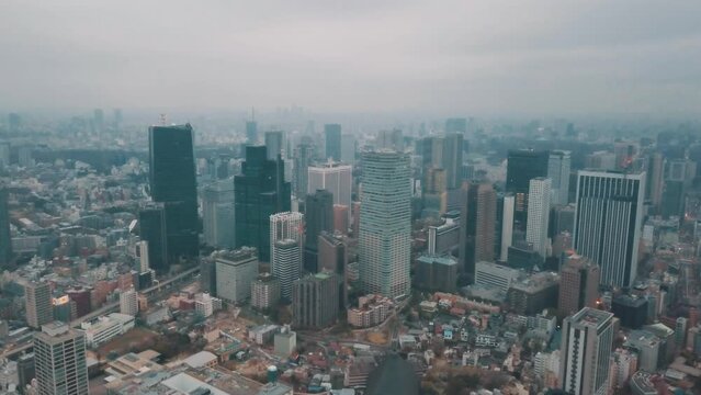 Aerial drone panning up away from the Tokyo city skyline in Japan on an overcast day with hazy smog filled skies and grey storm clouds above busy roads and skyscrapers