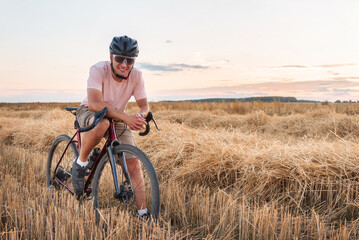 Fototapeta na wymiar Cyclist stands in the field at sunset. Beautiful landscape with man in helmet and sunglasses, gravel bicycle and dry grass. Sports lifestyle.