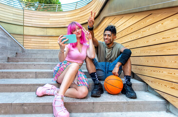 alternative diverse couple hanging out together sitting on city stairs making a selfie holding smartphone. interracial teenagers friends with basketball relaxing outdoors. tech and lifestyle concept