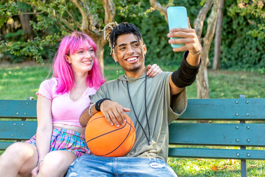 alternative diverse couple hanging out together sitting in a park bench making a selfie holding smartphone. interracial teenagers friends with basketball relaxing outdoors. tech and lifestyle concept