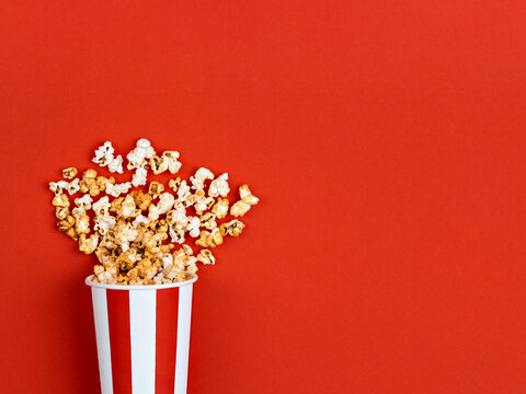 Popcorn spilled from a white and red striped bucket on a bright red background. Cinema, movies, movie night and entertainment concept. Top view, above, overhead.