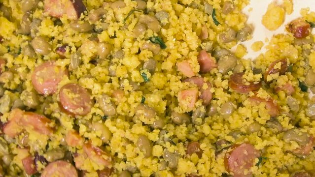 Cooking and mixing with a spoon the Northeastern Feijao Tropeiro, brazilian dish made with cuscuz, string bean, sausage, bacon, chive and curd cheese. Zoom out effect.