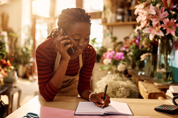 African American florist writing order while talking to customer over the phone at flower shop.