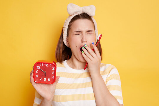 Image of sleepy young brunette woman wearing t shirt and hair band standing with red alarm clock and toothbrush, yawning, covering mouth with hand, posing isolated on yellow background.