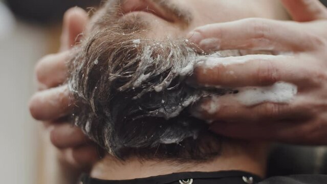 Hands of the master wash the beard of a hipster with shampoo, close-up. Barber washes man's beard before haircut