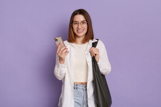 Image of adorable woman with black bag wearing white shirt and jeans posing isolated over purple background, using mobile phone, checking new app, looking smiling at camera.