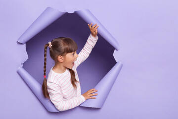 Horizontal shot of cute charming little girl with braids wearing striped shirt posing in torn paper wall, looking away with amazed expression, copy space for advertisement or promotion. - obrazy, fototapety, plakaty