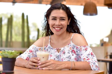 Mujer  feliz bonita latina sonriendo sentada en cafetería con bebida en  restaurante mediana edad 
