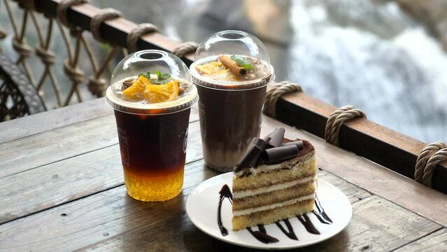 Plastic cups with fresh healthy cold drinks and plate with piece of organic vegan chocolate cake on wooden table in outdoor cafe with waterfall on background. Food and beverage for takeaway