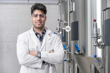 Young Handsome male worker in white lab coat smiling and looking to camera, standing at rows of steel brewing vats and crossing hands .Concept of brewery and beer plant. High quality photography.