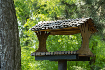 Close-up of wooden bird feeder on tree in green park. Handmade food house for birds with roof with round details. Bokeh effect blurred background.