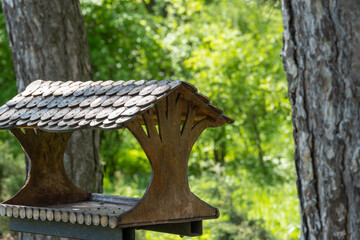 Close-up of wooden bird feeder on tree in green park. Handmade food house for birds with roof with round details. Bokeh effect blurred background.