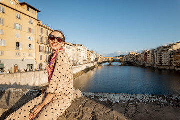 Fototapeta na wymiar Young woman enjoys beautiful view on famous Old bridge in Florence, sitting on the riverside at sunset. Female traveler visiting italian landmarks. Stylish woman wearing dress and colorful shawl