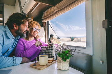 Fototapeta Tourist young adult people enjoy phone connection and freedom sitting inside a modern camper van and enjoying beach outside. Concept of summer holiday couple travel vacation planning next destination obraz