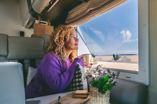 Serene adult woman admiring nature beach sitting inside a modern camper van in travel holiday vacation. Pretty young female people in van life enjoy coffee and breakfast morning parked free. Freedom