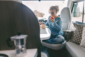 View of young woman sitting and having leisure time inside a camper van motor home. Healthy lifestyle female eating apple and using computer laptop. Alternative free off grid lifestyle - obrazy, fototapety, plakaty