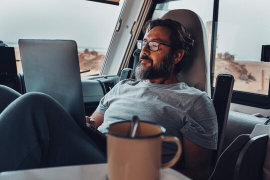 Digital nomad new modern job lifestyle with handsome adult man working and relaxing inside a camper van with beach and nature outside. Smart working free office concept eith laptop and connection