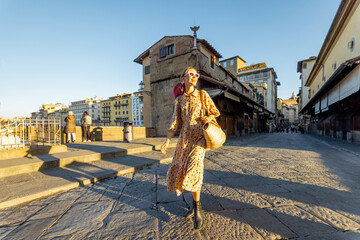 Woman walking on famous old bridge, called Ponte Vecchio, in Florence. Concept of traveling italian landmarks. Stylish woman with colorful shawl and sunglasses. Wide view on sunrise