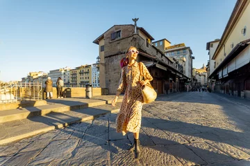 Acrylic prints Ponte Vecchio Woman walking on famous old bridge, called Ponte Vecchio, in Florence. Concept of traveling italian landmarks. Stylish woman with colorful shawl and sunglasses. Wide view on sunrise
