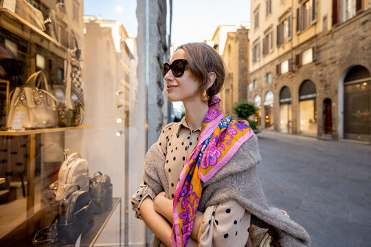 Fototapeta Beautiful woman looks on a showcase with bags and accessories of clothes shop in old town of in Florence city in Italy. Stylish woman wearing colorful shawl in retro italian style