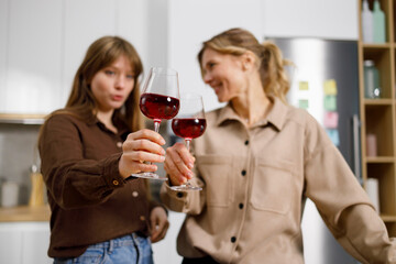 Mature woman and adult daughter clinking with glasses of red wine at home kitchen