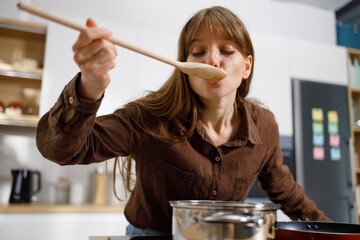 Young woman tasting food with wooden spoon while cooking