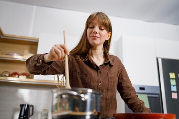 Young woman is stirring with a wooden spoon in a saucepan on induction stove. Housewife preparing...