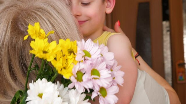  cute caucasian boy 7 years old with flowers hugs blonde mom, son gives mom a bouquet of white and yellow chrysanthemums, child love for mother, slow motion