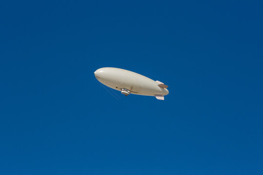 A white blimp without any markings, a blank canvas or banner space with a blue sky in the background. A lighter than air ship flying high with room to put your own ad.