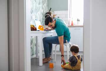 Father working on laptop while child playing on kitchen floor
