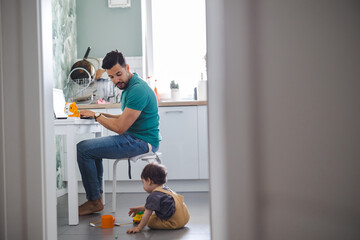 Father working on laptop while child playing on kitchen floor
