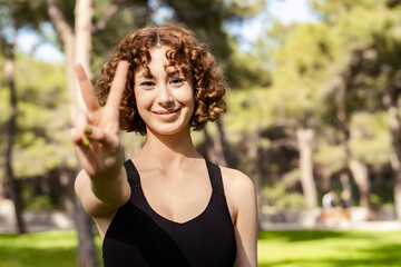 Young caucasian woman wearing black sports bra standing on city park, outdoor showing and pointing up with fingers number two while smiling confident and happy. Selective focus on her face.