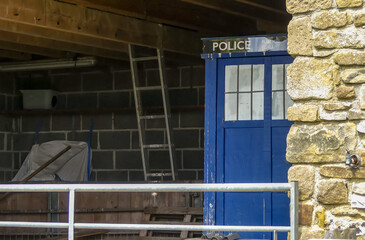Old unattended blue Police phone box which has landed in an agricultural building, with farming equipment, metal gate and ladder.  Iconic science fiction prop. Space for text. England. - 508852649