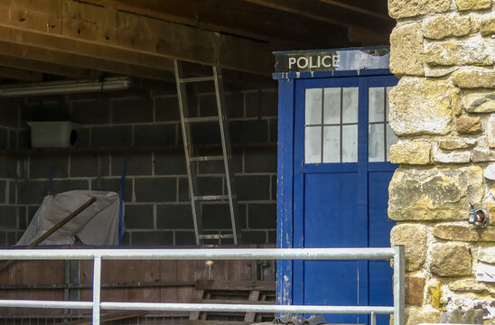 Old unattended blue Police phone box which has landed in an agricultural building, with farming equipment, metal gate and ladder.  Iconic science fiction prop. Space for text. England.