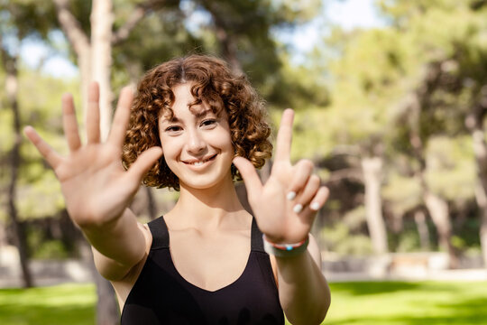Young caucasian woman wearing black sports bra standing on city park, outdoor showing and pointing up with fingers number seven while smiling confident and happy. Selective focus on her face.