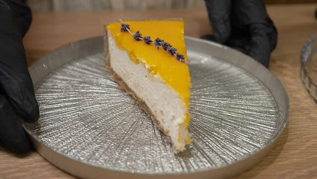 Female hands in black rubber gloves put metal plate with cheesecake on table. Piece of cheesecake with orange jam decorated with lavender branch closeup