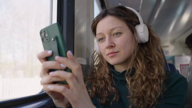 Young woman travels by electric train listening to music in white headphones. Curly woman in white headphones uses smartphone in train carriage