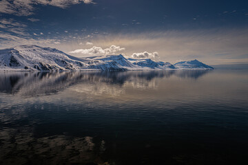Fototapeta na wymiar 2022-05-15 A ROW OF SNOW COVERED MOUNTAINS WITH A REFLECTION IN THE ARCTIC OCEAN AND NUMEROSU CLOUDS NEAR THE ISLAND OF SVALBARD NORWAY IN THE ARCTIC